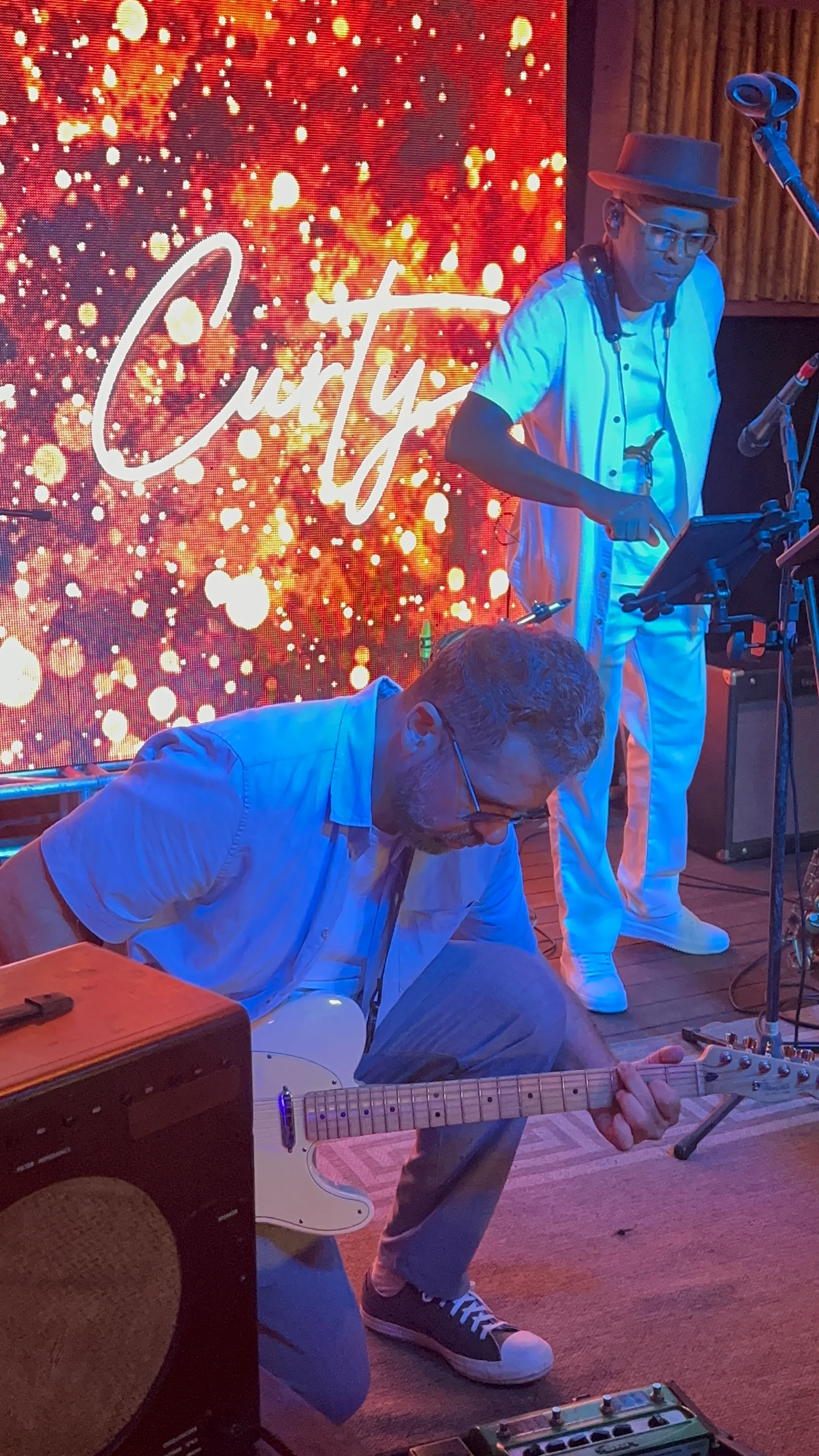 Pablo Curty ajoelhado tocando guitarra diante de um painel de luzes com o nome Curty.