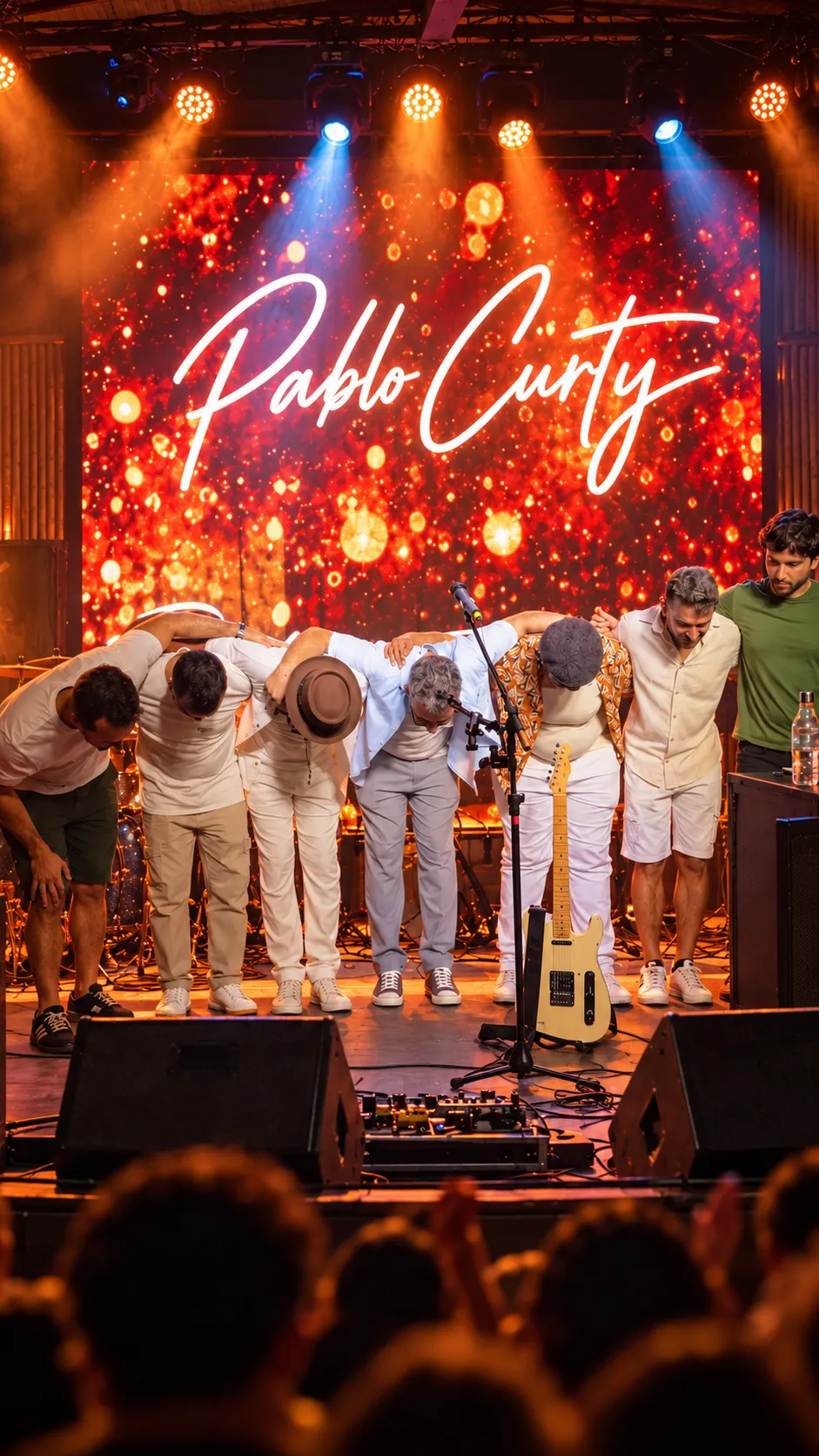 A banda abraçada agradecendo ao público, diante de um painel de luzes com o nome Pablo Curty.