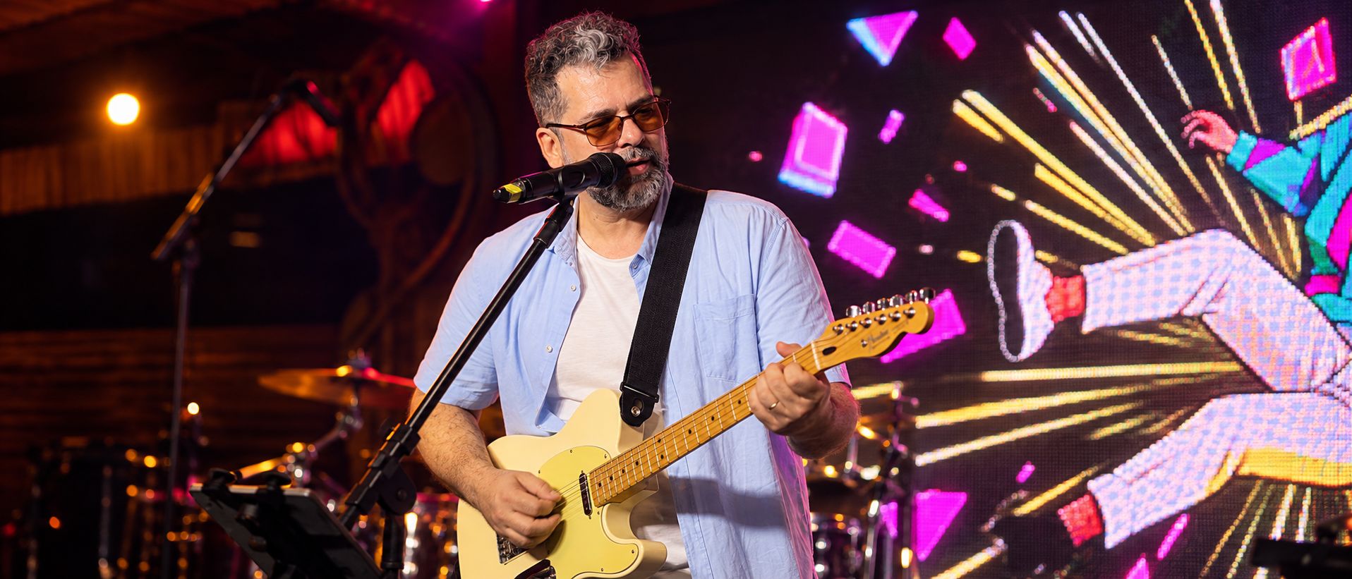 Pablo Curty cantando e tocando guitarra no palco, com a banda e um painel de luzes coloridas ao fundo.