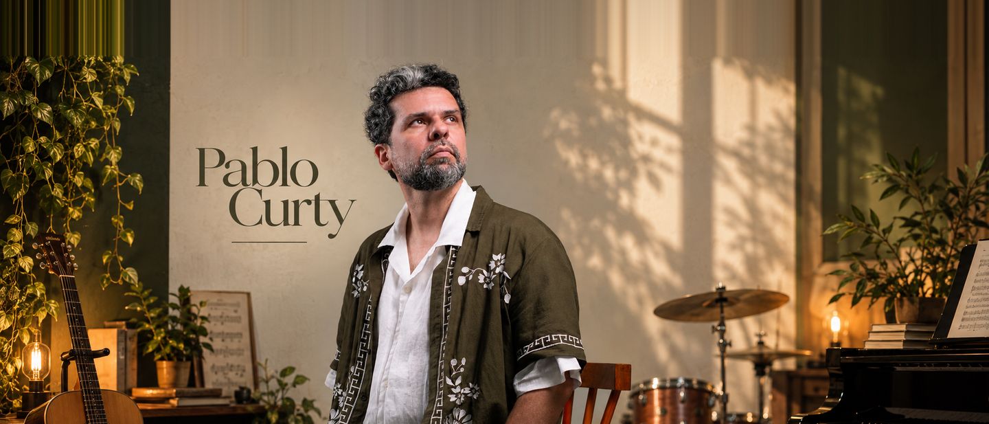 Pablo Curty de pé em um estúdio iluminado pelo sol da tarde, com violão, bateria e piano ao redor e o nome Pablo Curty escrito na parede.
